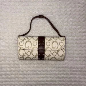 Beige och brun axelväska  - Snygg axelväska i beige med brunt mönster av loggor och detaljer i mörkbrunt skinn. Väskan har ett cylinderformat utseende, bred rem och dekorativa nitar framtill. Invändigt finns två fack med dragkedja för smart organisering.