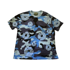 Blå Camo Denim Tears T-shirt  - Säljer en fet t-shirt från Denim Tears i storlek XL med blått och svart camouflage-mönster och coola vita blommor över hela. Rund hals och korta ärmar, tillverkad i mjuk bomull. Perfekt för dig som vill sticka ut med streetwear-vibe.