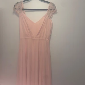 Ljusrosa mellan lång klänning från Boohoo - Superfin ljusrosa långklänning från Boohoo med v-ringning och spetsdetaljer på axlarna. Klänningen har smala axelband, lätt transparent tyg och dragkedja i ryggen. Perfekt för dig som vill ha en romantisk och flowy look.