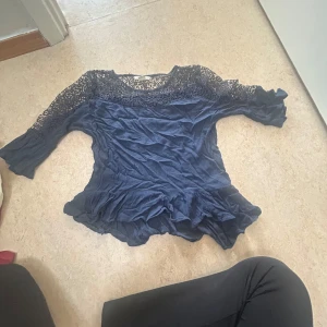 Mörkblå spetsblus från H&M - Säljer en mörkblå blus från H&M med vackra spetsdetaljer upptill och på ärmarna. Blusen har trekvartsärm, volangkant nertill och är tillverkad i ett lätt, flowy material. Perfekt för dig som gillar romantiska och feminina plagg.