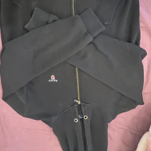 Svart hoodie från Gant med dragkedja - Svart hoodie från Gant med broderad logga på bröstet, dragkedja framtill och snörning i huvan. Klassisk modell med ribbade muddar och mjukt material, perfekt för chill dagar eller när du vill ha en clean look.