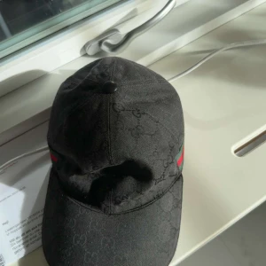 Svart Gucci keps med logomönster - Snygg svart keps från Gucci med diskret logomönster över hela ytan och klassiska gröna och röda ränder på sidan. Justerbar rem baktill och böjd skärm. Tillverkad i bomull med en cool streetstil som passar perfekt till din outfit.