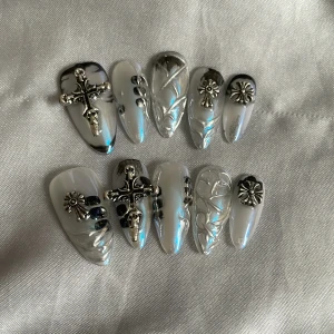 S - Chromed press-on naglar med kors och blommor - Snygga press-on naglar i kromad silverfärg med svart detalj, dekorerade med 3D-metallkors och blommor. Naglarna har en glansig, pärlemorskimrande finish och coola upphöjda mönster. Mix av mandel- och kistform, perfekt för dig som gillar edgy och unik nageldesign.