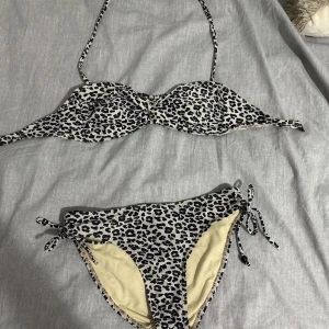 Leopardmönstrad bikini från Ocean Club - Snygg bikini från Ocean Club med leopardmönster i svart och beige. Överdelen har bygel och knytband i nacken, medan underdelen har knyt i sidorna för en justerbar passform. Perfekt för stranden eller poolen i sommar!