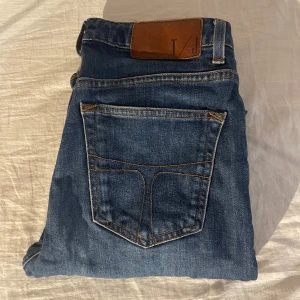 Blåa tiger of sweden jeans  - Snygga blå jeans från Tiger of Sweden med klassisk femficksdesign och bruna kontrastsömmar. Jeansen har rak passform och normal midja, tillverkade i slitstarkt denim med läderpatch bak i midjan. Perfekta för en stilren och avslappnad look.