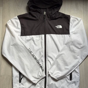 Svartvit vindjacka från The North Face - Snygg svartvit vindjacka från The North Face med huva och hel dragkedja. Jackan har två fickor med dragkedja och logga på bröstet samt längs ena ärmen. Lätt och smidig modell i syntetmaterial, perfekt för blåsiga dagar.