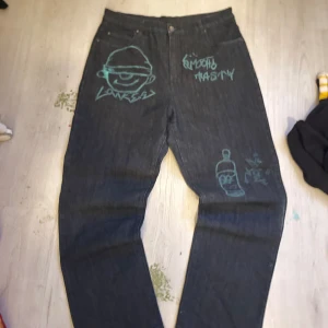 Svarta jeans från Tasty med graffiti - Unika svarta jeans från Tasty med graffiti-inspirerade turkosa teckningar och text på framsidan. Klassisk femficksmodell med raka ben och broderad detalj på bakfickan. Jeansen har normal passform och är tillverkade i slitstarkt denimtyg.