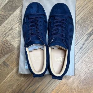 Mörkblå sneakers från Christian Louboutin - Snygga mörkblå sneakers i mocka från Christian Louboutin med klassisk röd sula och diskret logga på sidan. Skorna har rund tå, platt sula och snörning. Perfekta för dig som vill ha en lyxig och stilren look med en unik touch.