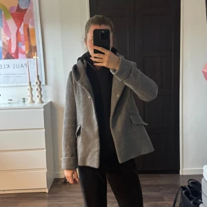 Grå ullkappa från Zara XS - Säljer en stilren grå kappa från Zara i storlek XS. Jackan har klassisk krage, snygga fickor med lock och är tillverkad i ullblandning. Perfekt för dig som gillar en clean och tidlös look. Fodrad insida och uppvikta ärmslut. Har använts och finns defekter!