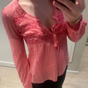 Rosa blus med broderi och knyt - Supersöt rosa blus med broderade detaljer över bysten och knyt framtill. Långärmad modell med lätt v-ringning och volangkant vid ärmslut och hals. Tunn och luftig i bomull, perfekt för vår och sommar.