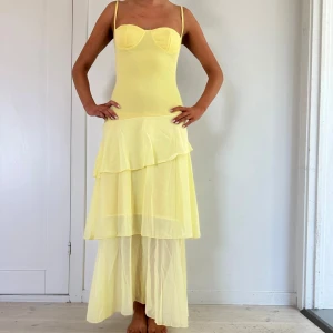 Ljusgul långklänning med volanger - Säljer en ljusgul långklänning med smala axelband och figurnära överdel. Kjolen har flera lager med volanger och är tillverkad i ett lätt, transparent material som ger en luftig känsla. Perfekt för sommarens tillfällen!