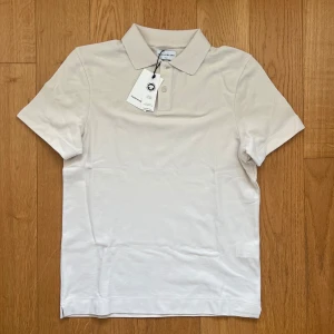 Beige pikétröja från These Glory Days - Snygg beige pikétröja från These Glory Days med klassisk krage och två knappar framtill. Tillverkad i mjuk bomull som känns skön mot huden. Perfekt för en clean och stilren look. Kortärmad modell med normal passform. Skriv om ni har frågor eller vill diskutera pris