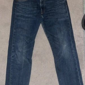 Levi's 501 jeans blå W30 L30 - Klassiska Levi's 501 jeans i blå denim med raka ben och normal passform. Byxorna har fem fickor, knappgylf och snygga kontrastsömmar. Märket syns tydligt på bakfickan och läderpatchen i midjan. Perfekta för en avslappnad och tidlös look.
