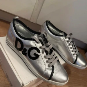 Silvriga sneakers från D&G - Snygga sneakers från Dolce & Gabbana i metallic-silver med svarta detaljer och D&G-logga på sidan. Skorna har snörning, svart kant runt öppningen och platt sula. Perfekta för dig som vill sticka ut med din stil. Skorna är köpa i  NK Stockholm