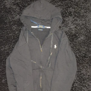 Svart hoodie från Polo Ralph Lauren - Svart hoodie från Polo Ralph Lauren med dragkedja och huva. Klassisk vit logga broderad på bröstet och snörning i huvan. Tillverkad i mjuk bomull, perfekt för chill dagar. Långa ärmar och känguruficka framtill.