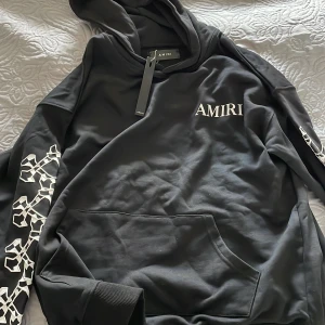 Svart hoodie från Amiri med tryck - Svart hoodie från Amiri i 100% bomull med vit logga på bröstet och coolt grafiskt tryck längs ärmarna. Klassisk känguruficka framtill, ribbade muddar och huva. Snygg detalj med metallbricka med Amiri-logga. Perfekt för dig som gillar streetwear Den e även helt ny.