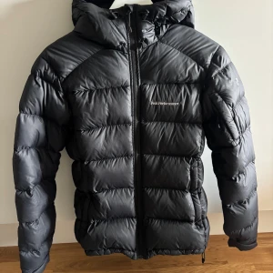Svart dunjacka Peak Performance  - Svart dunjacka från Peak Performance med huva och dragkedja framtill. Jackan är quiltad och har en clean, sportig look med diskret logga på bröstet. Perfekt för kalla dagar och riktigt skön att bära. Klassisk passform och snygg design.