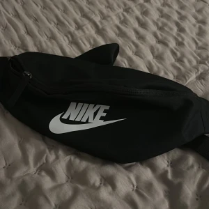 Svart magväska från Nike - Säljer en svart magväska från Nike med klassisk vit logga framtill. Väskan har justerbart band och dragkedja upptill. Perfekt för att bära mobilen, plånboken och andra smågrejer när du är på språng.