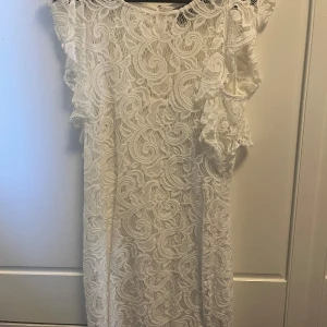 Vit spetsklänning från Vero Moda - Superfin vit spetsklänning från Vero Moda med vackra volangdetaljer på axlarna. Klänningen har ett snyggt spetsmönster över hela och är ärmlös. Perfekt för sommarens alla tillfällen och ger en romantisk vibe.