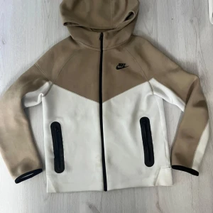  beige nike tech barn 147-158 - Snygg vindjacka från Nike i beige och vitt med svart logga på bröstet. Jackan har huva, hel dragkedja och två svarta fickor med dragkedja framtill. Perfekt för dig som gillar sportig stil och vill ha en jacka som sticker ut med sin coola färgblock-design.