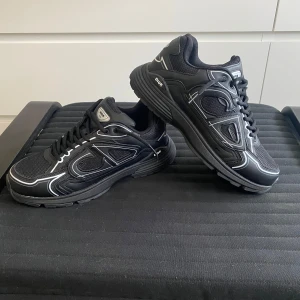 Skor - Säljer ett par svarta sneakers med vita kontrastdetaljer och meshpaneler. Skorna har rund tå, snörning och tjock sula för en sportig vibe. Märkeslogga syns både fram och bak. Perfekta för dig som vill sticka ut med lyxig streetstyle. Låda medföljer tyvärr inte! 