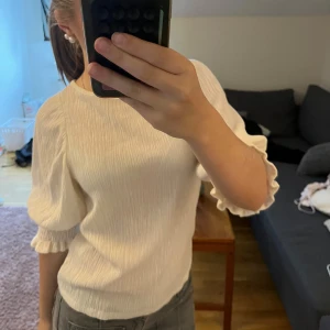 Krämvit blus med puffärm och struktur - Säljer en krämvit blus med puffiga trekvartsärmar och volangdetaljer vid ärmslut och midja. Toppen har en snygg strukturerad yta och är lätt och luftig, perfekt för vår och sommar. Passformen är normal och materialet känns mjukt mot huden.