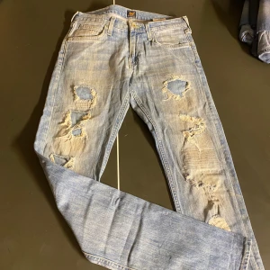 Ljusblå slitna jeans från Lee - Säljer ett par ljusblå jeans från Lee med slitna detaljer och hål på både fram- och baksida. Klassisk femficksmodell med Lee-lapp bak i brunt läder. Jeansen har en rak passform och är tillverkade i bomullsdenim.
