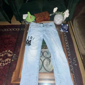 Ljusblå Amiri jeans med logga Waist 30 length 31 - Säljer ett par ljusblå jeans från Amiri med svart tryckt logga och metallmärke på benet. Jeansen har klassisk femficksdesign, raka ben och är tillverkade i bomullsdenim. Perfekta för dig som vill sticka ut med streetwear-vibe.