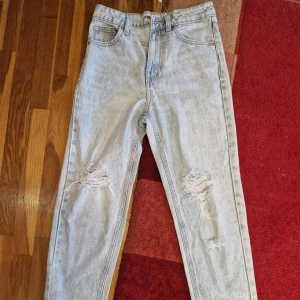 Ljusblå slitna jeans med hög midja - Säljer ett par ljusblå jeans med snygga slitningar på båda knäna. Jeansen har hög midja och klassisk femficksdesign. Perfekta för dig som gillar en avslappnad och trendig stil.