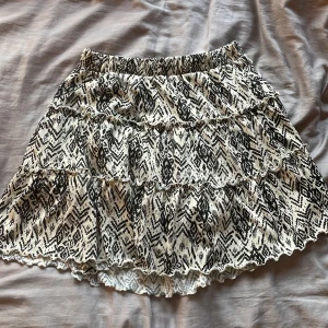 Svartvit mönstrad kjol från Lindex - Kort kjol från Lindex i svartvitt mönster med volangdetaljer och resår i midjan. Kjolen har ett coolt grafiskt print och är gjord i ett mjukt, lätt material. Perfekt för dig som gillar en avslappnad men trendig stil. Med insydda shorts!! 