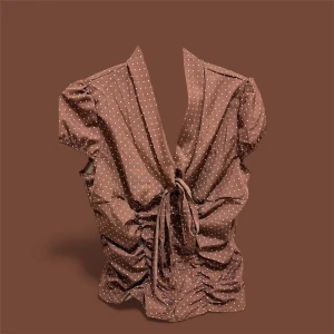 Brun prickig omlottblus med knyt - Supersöt brun blus med vita prickar och omlottmodell. Djup v-ringning framtill, knyt i midjan och rynkade detaljer längs sidorna. Korta puffärmar med resår. Perfekt för dig som gillar vintagevibbar och lekfulla mönster.