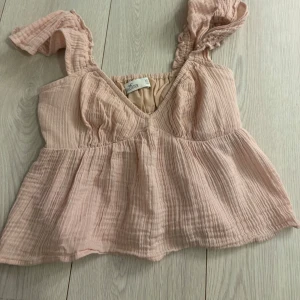 Snygg Festopp🤍 - Supersöt ljusrosa linnetopp från Hollister med volangig och luftig passform. Toppen har breda axelband med rynkade detaljer och v-ringning. Tillverkad i mjuk bomull med struktur för en chill och somrig vibe. Knappast använd.