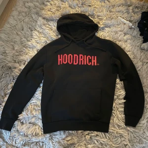 Svart hoodie från Hoodrich med röd logga - Svart hoodie från Hoodrich i storlek medium med stor röd logga på bröstet. Klassisk känguruficka framtill, huva med snörning och mjukt material som känns skönt mot huden. Perfekt för dig som gillar streetwear och vill sticka ut med en fet logga.