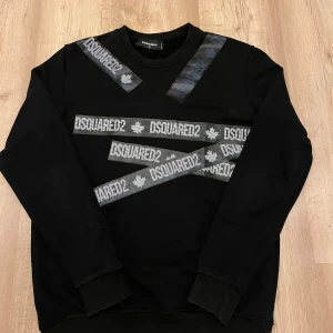 Svart sweatshirt från Dsquared2 - Cool svart sweatshirt från Dsquared2 med flera breda band med logga och lönnlövsmotiv tvärs över framsidan. Tröjan har rund halsringning och långa ärmar. Perfekt för dig som gillar streetwear och vill sticka ut med en unik design. Storleken är M men sitter som S, pris går att diskuteras då den har en liten fläck på tröjan men den borde försvinna i tvätten 