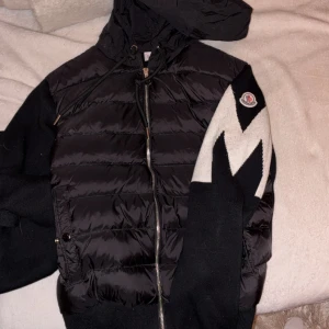 Svart Moncler pufferjacka med mönstrad ärm - Svart pufferjacka från Moncler med huva och dragkedja framtill. Ena ärmen har ett stort vitt och svart zigzag-mönster samt Moncler-logga. Jackan har snörning vid huvan och ribbade muddar. Perfekt för dig som gillar streetstyle och vill sticka ut.