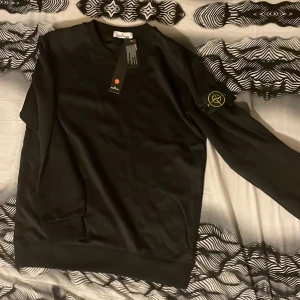 Svart sweatshirt från Stone Island - Svart sweatshirt från Stone Island med klassisk rund hals och ikonisk logotyp-patch på vänster ärm. Tröjan har långa ärmar och ribbade muddar vid ärmslut och nederkant. Perfekt för en clean och stilren look.