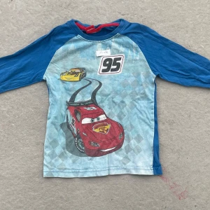 Blå Cars långärmad tröja Disney - Långärmad tröja från Disney Pixar med motiv av Blixten McQueen och Cruz Ramirez från Cars. Tröjan har blå ärmar, ljusblå framsida med rutigt mönster och tryck med nummer 95. Perfekt för alla små Cars-fans!