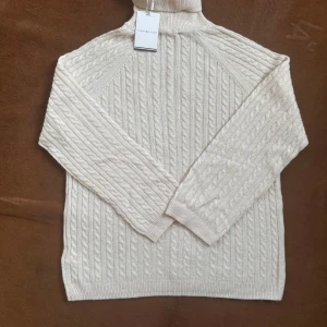 Vit polotröja från Tommy Hilfiger - Kabelstickad vit polotröja från Tommy Hilfiger med klassisk design. Tröjan har lång ärm, ribbade muddar och hög polokrage. Perfekt för kyliga dagar och enkel att styla med jeans eller kjol.