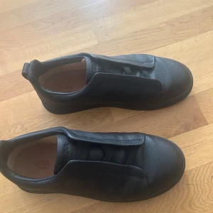 Svarta sneakers från Zegna i skinn - Snygga svarta sneakers från Zegna i mjukt, strukturerat skinn. Skorna har en stilren design med dold snörning och rund tå. Perfekta för dig som gillar minimalistisk och clean stil. Passar till det mesta och ger en lyxig känsla till outfiten.