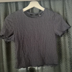 Grå crinkle topp från Vero Moda - Supersnygg grå topp från Vero Moda i crinkle-material. Modellen har korta ärmar med lätt volangkant och rund halsringning. 