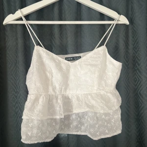 Vit volangig linnetopp från SHEIN - Supersöt vit linnetopp från SHEIN i storlek XS. Toppen har tunna axelband, broderade blommor och dubbla volanger för en luftig känsla. Dragkedja i sidan för enkel påklädning. Perfekt för varma dagar och somriga outfits!