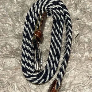 Randigt flätat bälte med läderdetaljer - Snyggt flätat bälte i blått och vitt med randigt mönster. Bältet har bruna läderdetaljer vid spännet och änden, samt ett klassiskt metallspänne. Perfekt accessoar för att lyfta din outfit med en stilren touch.