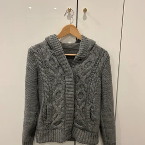 Grå stickad kofta med huva - Mysig grå stickad kofta med huva och grovt flätmönster framtill. Koftan har ribbade muddar och bred krage, samt två fickor på sidorna. Perfekt för kyliga dagar och enkel att matcha med det mesta.