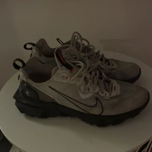 Nike React Element 55 sneakers vita - Nike React Element 55 sneakers i vitt med svarta detaljer och orange accenter. Skorna har en chunky svart sula, snörning och en sportig design. Ovandelen är i syntetmaterial med tydliga sömmar och Nike-logga på sidan. Perfekta för dig som gillar streetwear och bekväma sneakers.