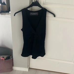 Svart omlott topp från Zara Collection - Svart topp från Zara Collection i omlottmodell med v-ringning och knyt framtill. Västen har en lätt utsvängd nederkant och är tillverkad i ett mjukt, slätt material som ger en stilren look. Perfekt att styla med jeans eller kjol för en trendig vibe.