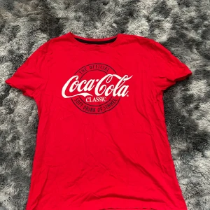 Röd Coca-Cola Classic t-shirt - Röd t-shirt från Coca-Cola med klassisk logga i vitt och svart tryck på bröstet. Rund halsringning och korta ärmar. Tillverkad i mjuk bomull, perfekt för dig som gillar retro och streetstyle.