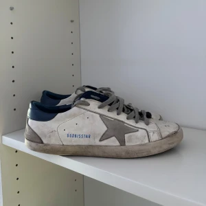 Golden Goose Superstars - Golden Goose Superstars i vitt med grå mockadetaljer och ikonisk stjärna på sidan. Skorna har grå snörning, blå hälkappa och låg profil. Perfekta för dig som gillar en avslappnad och trendig streetstyle. OBS! Större defekter, säljer billigt därmed. Hör av vid bild på defekt osv! 👍🏼