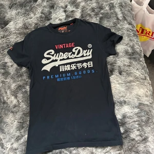 Svart Superdry t-shirt med tryck - Svart t-shirt från Superdry med stort vitt och blått tryck på bröstet och japanska tecken. Klassisk rund hals och korta ärmar. Skön passform och tillverkad i mjuk bomull. Perfekt för dig som gillar streetstyle och tydliga loggor.