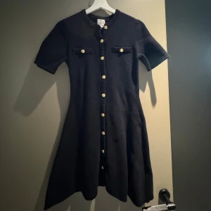 Svart stickad kortklänning med knappar - 100% cotton Supersnygg svart stickad klänning med korta ärmar och guldiga knappar hela vägen framtill. Klänningen har två dekorativa fickor på bröstet med knappar och ribbade detaljer längs kanterna. 