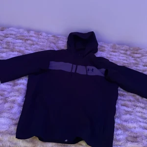 Svart jacka från Under Armour  - Svart jacka från Under Armour med grått band och logga över bröstet. Jackan har huva med snörning, lång ärm och dragkedja framtill. Modellen är loose fit och perfekt för chill eller träning. Materialet är mjukt och andas bra.